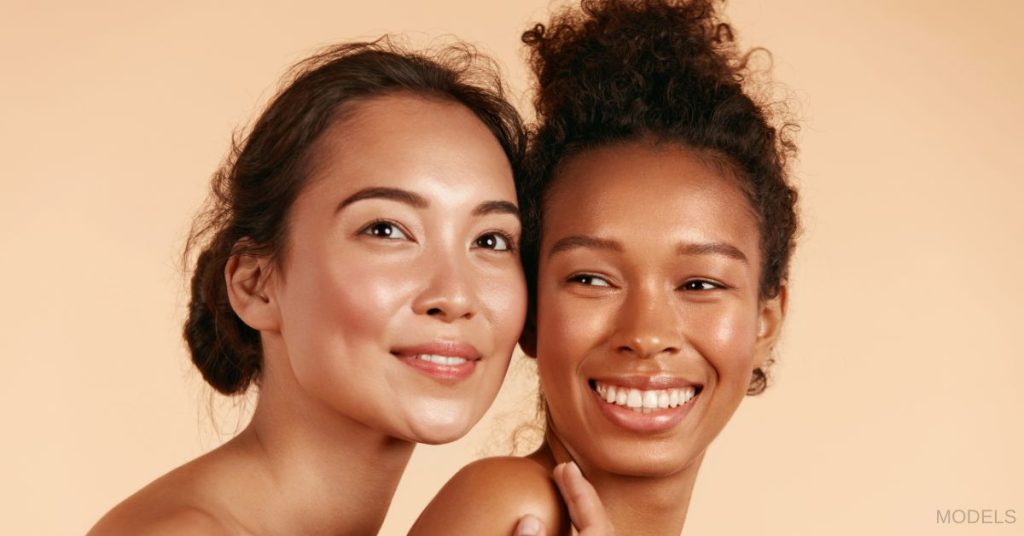 Two women with glowing skin smiling and looking outward. (MODELS)