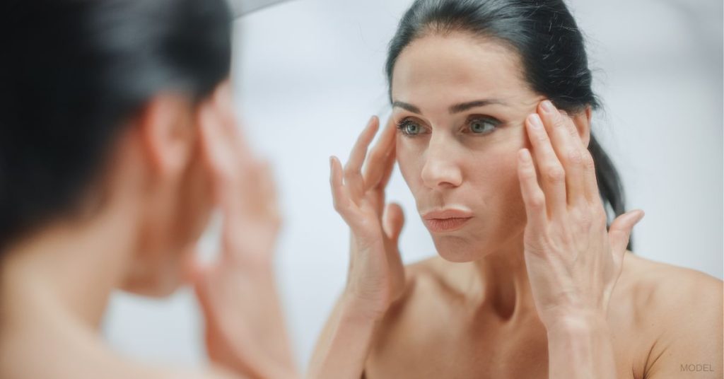 Woman looking in mirror and holding back her face by her temples. (MODEL)