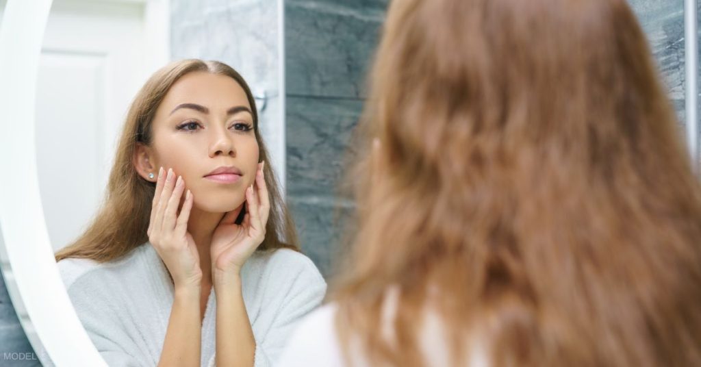 Woman looking in mirror and holding her face at the jawline. (MODEL)