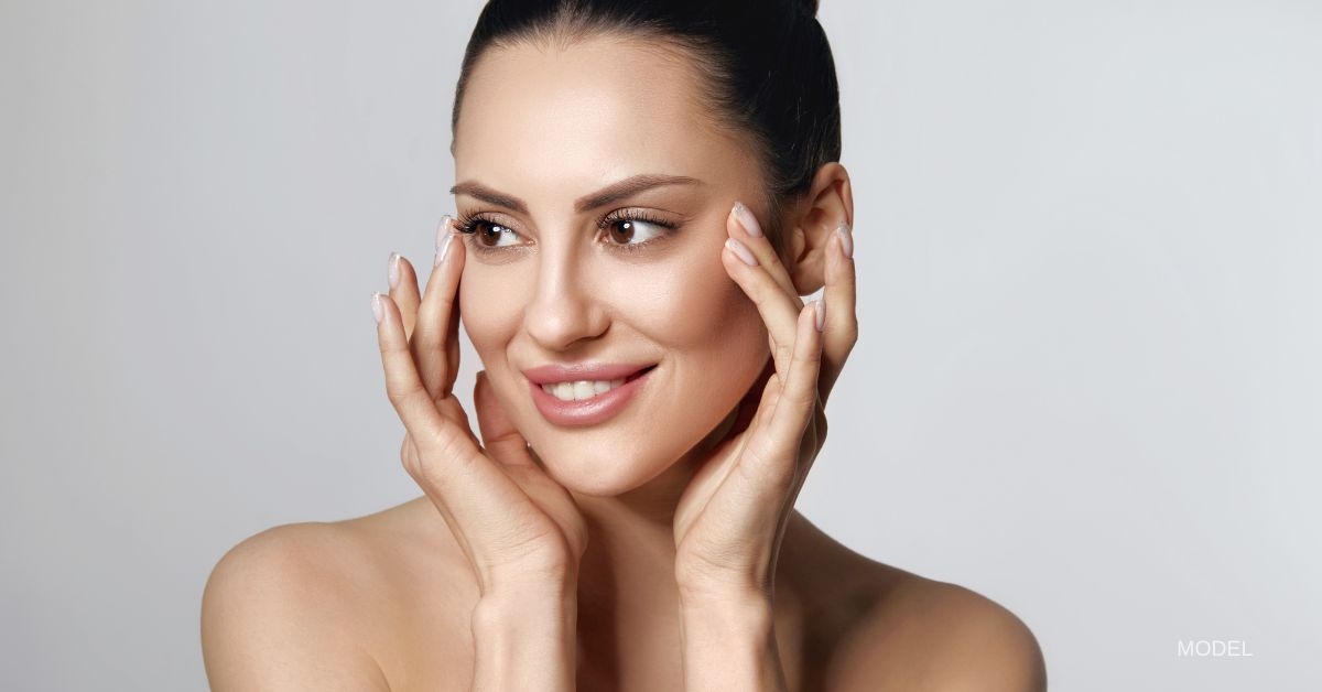 Woman with glowing skin, smiles while touching her face. (Model)